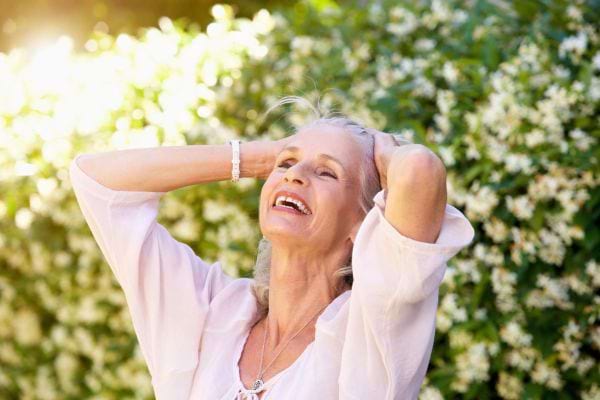 Femme senior souriante, mains dans les cheveux, profitant d’un moment de joie au jardin.