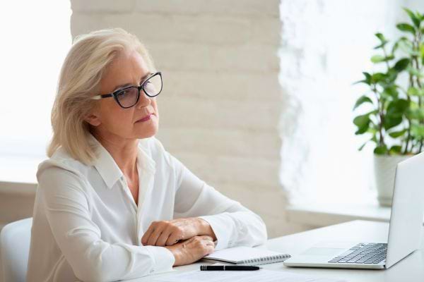 Femme senior aux cheveux blancs assise devant un ordinateur, l’air pensive.