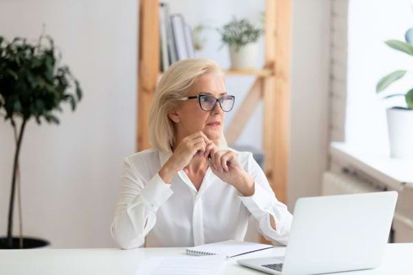 Femme âgée assise au bureau, regardant par la fenêtre d’un air pensif.