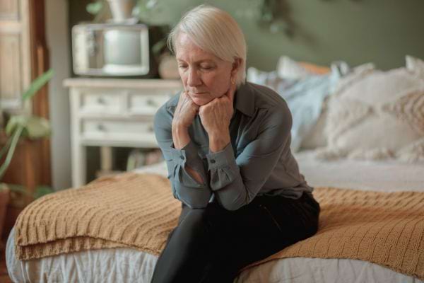 Femme senior assise sur un lit, tête baissée, semblant préoccupée.
