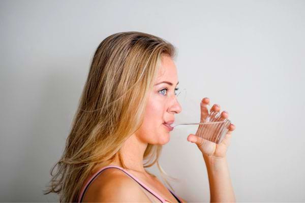 Femme blonde buvant un verre d’eau claire contre un mur blanc.