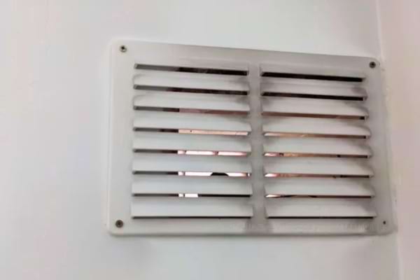 Une grille de ventilation blanche légèrement poussiéreuse est fixée sur un mur.