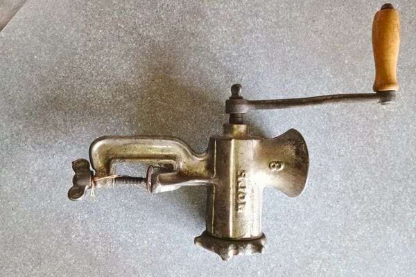 Ancien hachoir à viande en métal avec manivelle en bois posé sur une table.