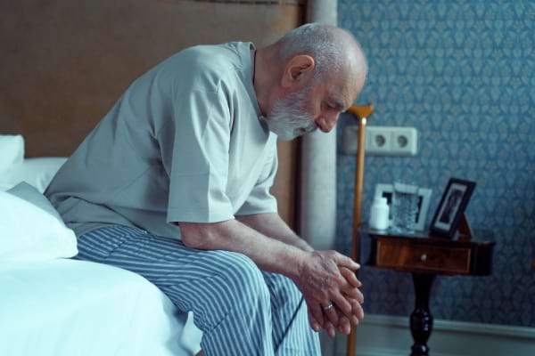 Un homme âgé assis sur son lit, pensif, les mains jointes et la tête baissée.