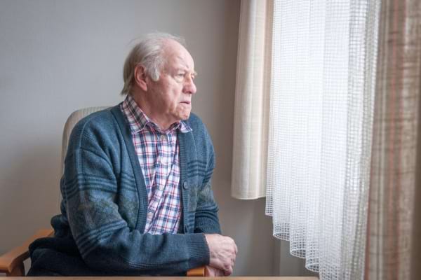 Un homme âgé en cardigan bleu regarde pensivement par la fenêtre.