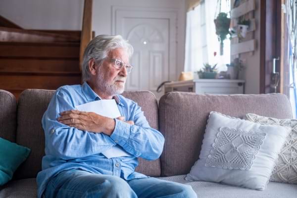Un homme âgé assis sur un canapé tient une lettre contre lui, le regard triste.