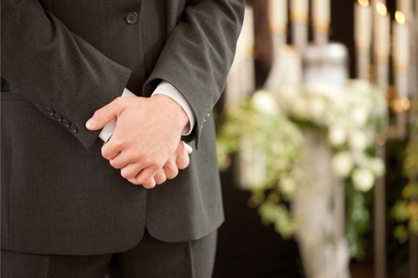 Homme en costume tenant ses mains devant un cercueil fleuri.