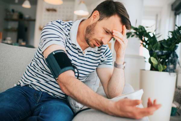 Un homme assis sur un canapé, fatigué et inquiet, mesure sa tension artérielle avec un tensiomètre.