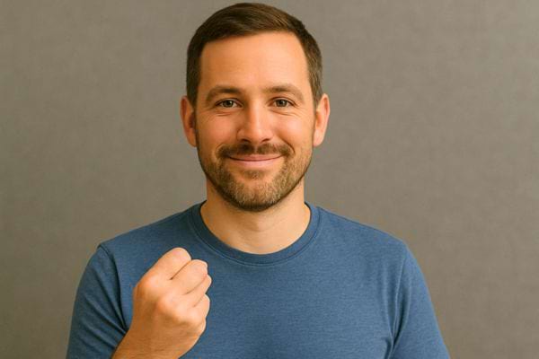 Un homme barbu en t-shirt bleu sourit en levant légèrement la main fermée.