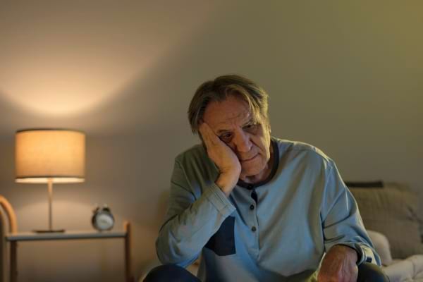 Un homme plus âgé, en pyjama, assis sur son lit, l’air fatigué et mélancolique.