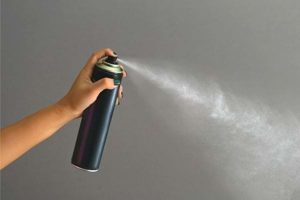Main tenant une bombe aérosol vaporisant un produit sur fond gris neutre.