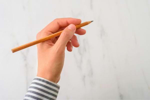 Main tenant un crayon en bois sur fond clair, vêtue d’une chemise à rayures.