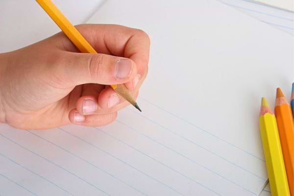 Main tenant un crayon jaune écrivant sur une feuille lignée avec crayons colorés posés à côté.