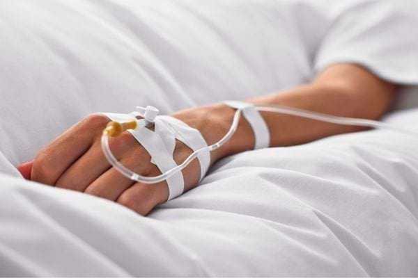 Une main avec perfusion repose sur un drap blanc dans un lit d’hôpital.