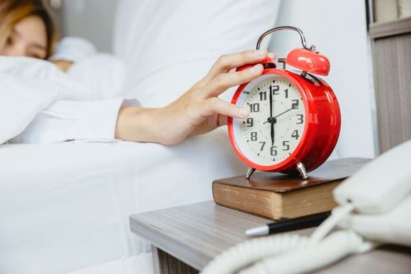 Une main tendue arrête un réveil rouge posé sur une table de chevet.