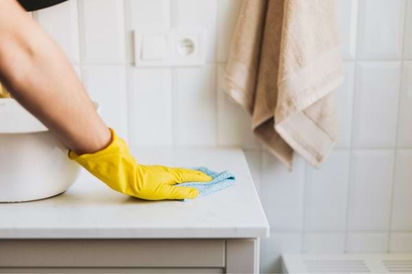 Main avec gant jaune essuyant une surface blanche dans une salle de bain carrelée.