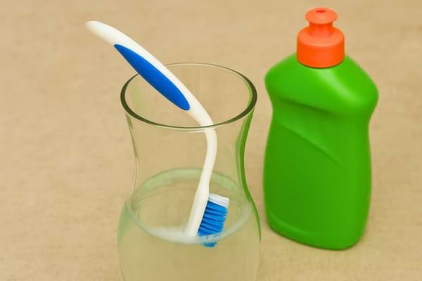 Une brosse à dents trempe dans un verre d’eau savonneuse à côté d’un produit vaisselle.