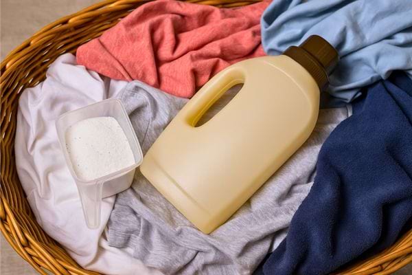 Panier en osier contenant du linge coloré, un bidon de lessive et une dose de poudre.