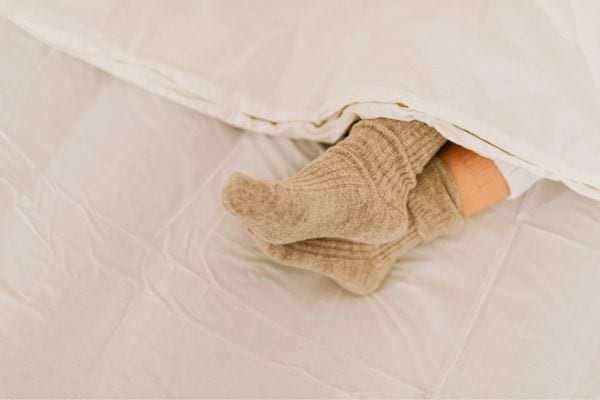 Pieds en chaussettes beiges dépassant d’une couette blanche sur un lit.