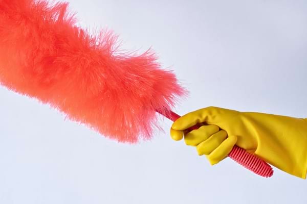 Une main gantée de jaune tient un plumeau orange pour dépoussiérer.