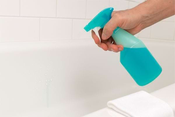 Une main vaporise un produit nettoyant bleu sur la surface d’une baignoire.