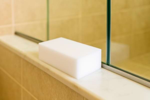 éponge à mélamine blanche posée sur le rebord d’une douche en verre