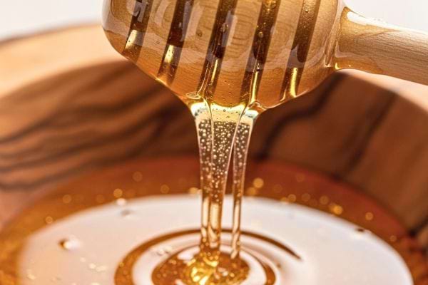 Du miel doré s’écoule d’une cuillère en bois dans un bol.