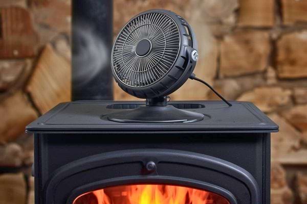 Petit ventilateur électrique noir posé sur un poêle à bois allumé.