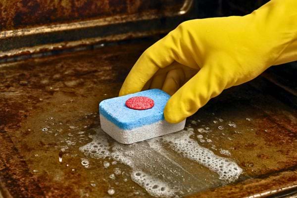 Une main gantée frotte l’interieur sale du four avec une tablette lave-vaisselle pour enlever la graisse incrustee.