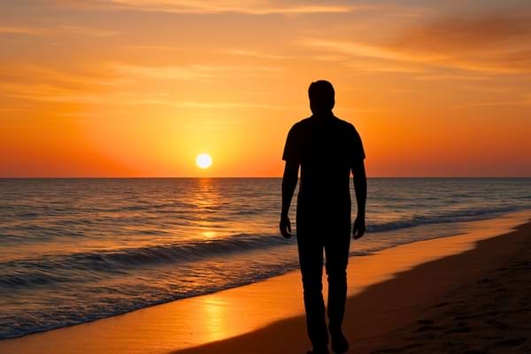 A man walking alone on a peaceful beach at sunset, silhouette against the light,