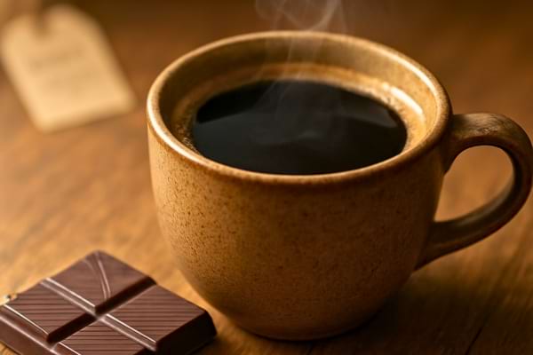 une tasse de caf&eacute; fumante et un carr&eacute; de chocolat noir pos&eacute;s sur une table en bois rustique