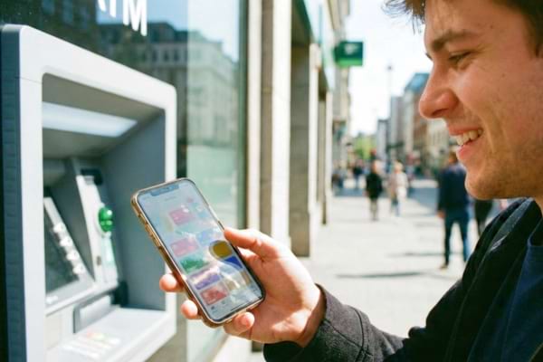 Un homme jeune, l'air soulag&eacute;, tient son smartphone devant un distributeur automatique de billets