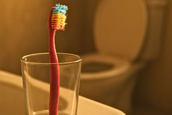 une brosse &agrave; dents color&eacute;e pos&eacute;e dans un verre sur le rebord d'un lavabo. En arri&egrave;re-plan flou, on devine des toilettes 