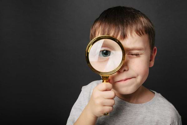 un enfant regarde a travers une loupe en fermant un oeil sur fond sombre