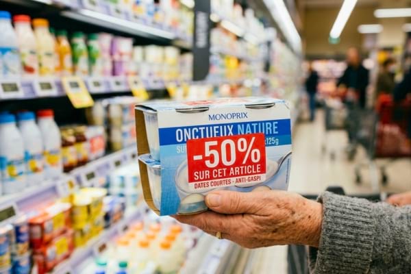 une main de senior tenant un paquet de yaourts avec une grosse étiquette de réduction rouge "-50%"