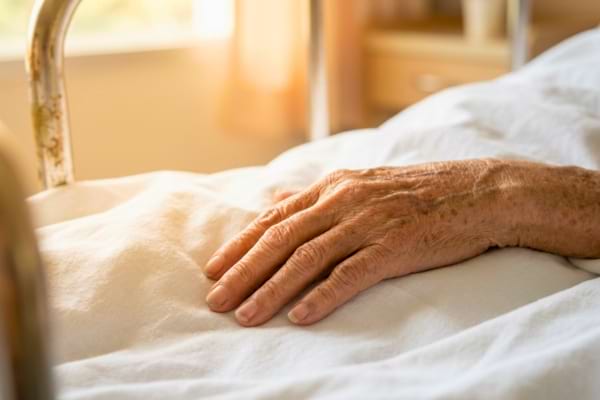 une main âgée et ridée reposant sur des draps blancs d'hôpital.