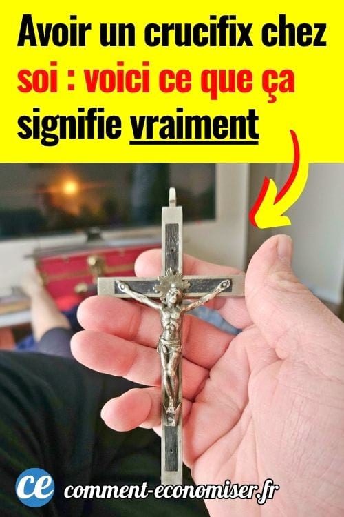 Main tenant un crucifix en m&eacute;tal devant un salon flou avec t&eacute;l&eacute;vision en arri&egrave;re-plan.