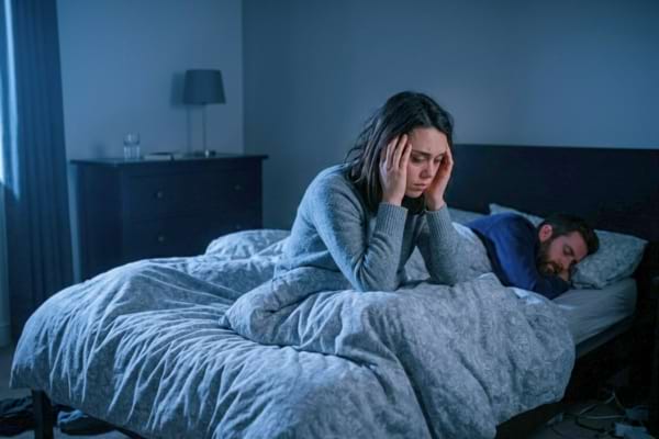 Une femme est réveillée, assise dans le lit, tenant sa tête dans ses mains, l'air épuisé et anxieux