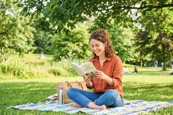 Une personne souriante en train de lire un livre dans un parc ensoleill&eacute;,
