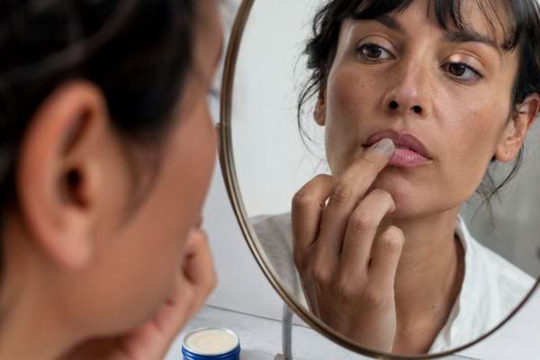 Femme appliquant une cr&egrave;me sur ses l&egrave;vres s&egrave;ches devant un miroir de salle de bain.