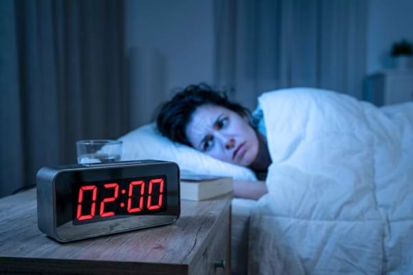 un r&eacute;veil digital affichant 02:00 pos&eacute; sur une table de chevet en bois. En arri&egrave;re-plan flou, une personne sous une couette