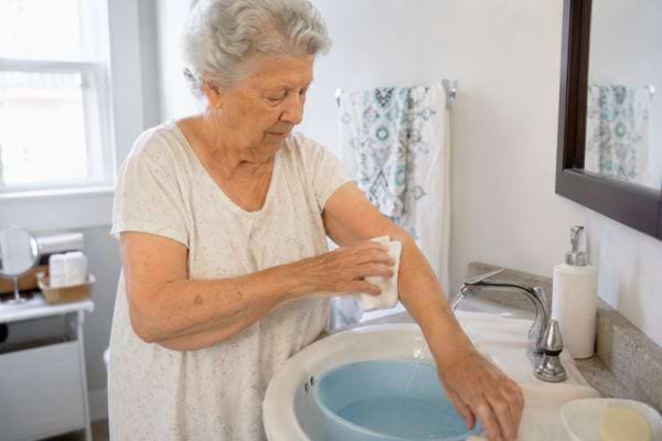Femme &acirc;g&eacute;e se lavant le bras au gant pr&egrave;s du lavabo.