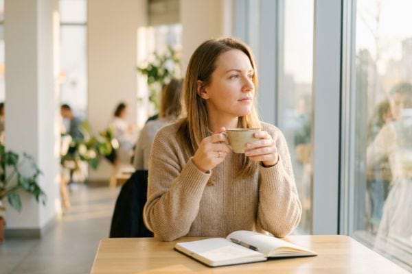 Femme seule buvant un caf&eacute; pr&egrave;s d&rsquo;une fen&ecirc;tre avec un carnet ouvert
