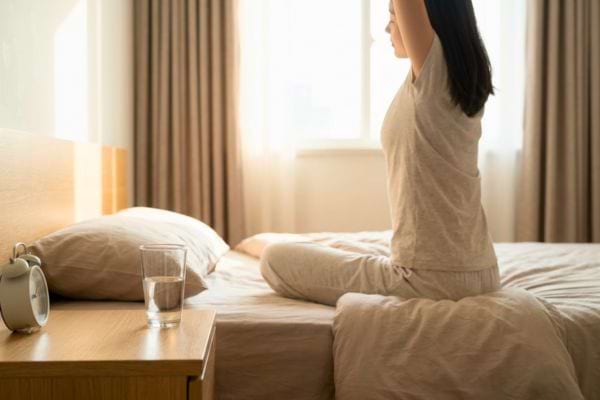 Femme assise sur un lit s’étirant au réveil avec un verre d’eau à côté