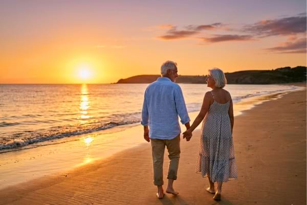 un couple de seniors souriants vus de dos, marchant sur une plage au coucher du soleil