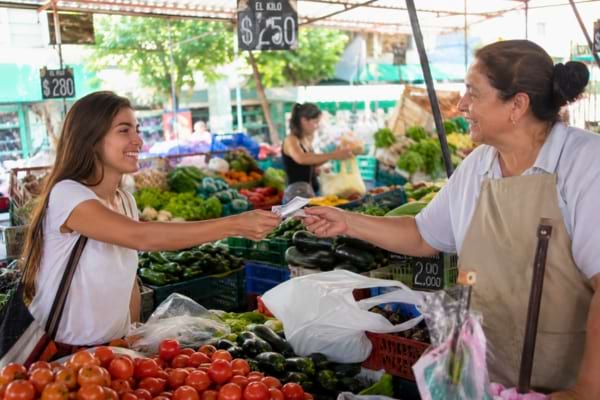 un jeune adulte souriant tendant un billet de banque à un commerçant sur un marché