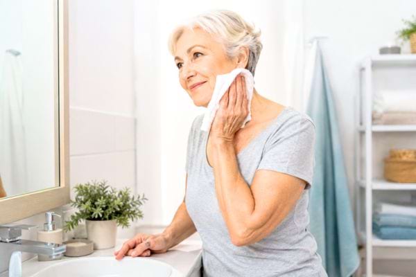 Femme senior faisant une toilette douce au gant devant le lavabo, dans une salle de bain propre et claire.