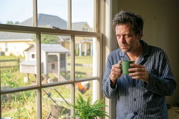 un homme fatigué en pyjama, tenant une tasse de café près d'une fenêtre, regardant avec exaspération vers le jardin