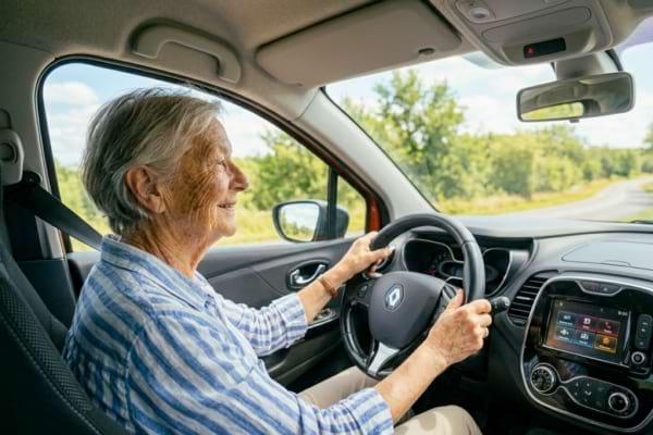 une personne de 80 ans souriante au volant de sa voiture par une journée ensoleillée