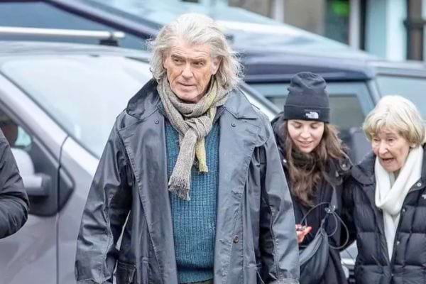Portrait de Pierce Brosnan aux cheveux longs gris, en manteau noir, devant une voiture claire